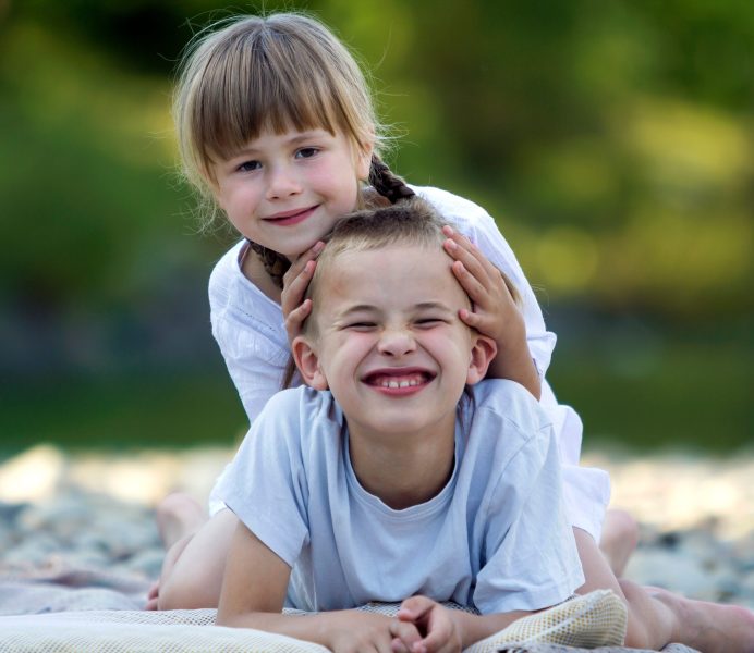Two young happy cute blond smiling children, boy and girl, brother and sister laying embraced on pebbled beach on blurred bright sunny summer day background. Friendship and perfect holiday concept.