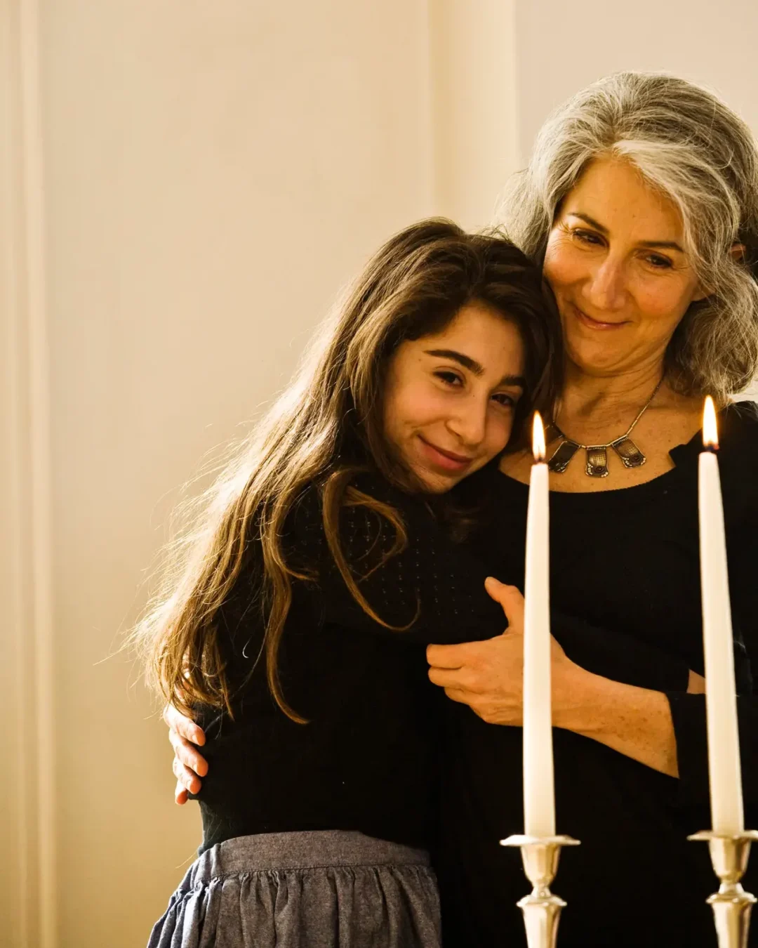 Two women, an older woman and a younger girl, embrace beside lit candles in a warm, intimate moment.
