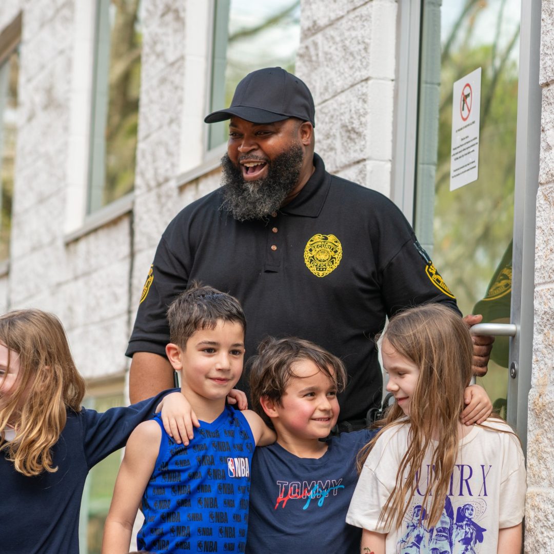Smiling security guard in a black polo and cap posing with a group of children outside a building.