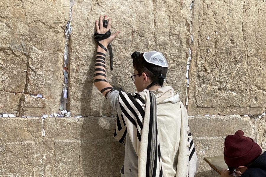 Person at the Western Wall wearing a tallit and tefillin, pressing a hand to the stones with leather straps wrapped around the arm. scattered prayer notes lie at the base of the wall.