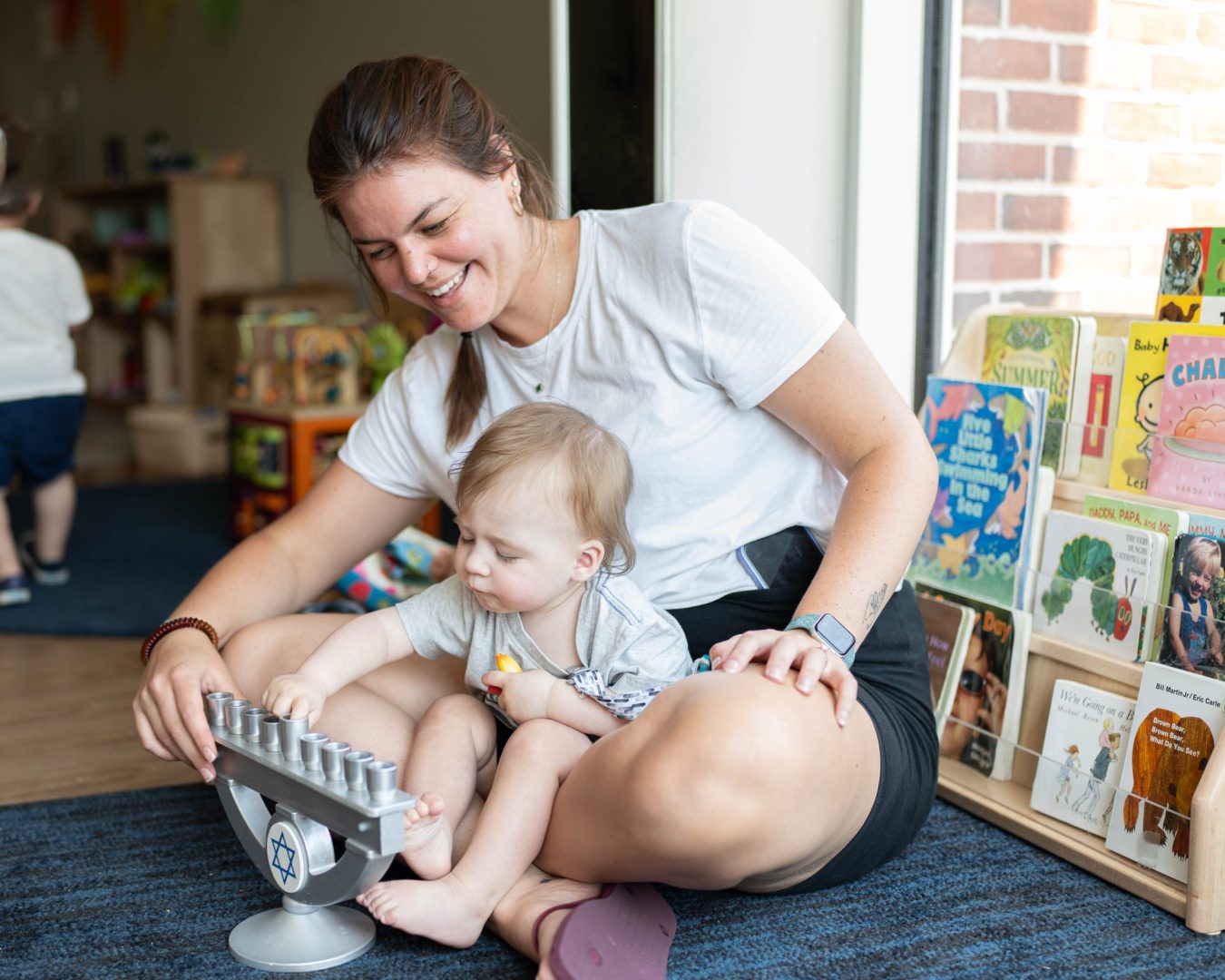 Mother and toddler sitting on a blue carpet in a kids' library, playing with a metal counting scale while shelves of books stand nearby.