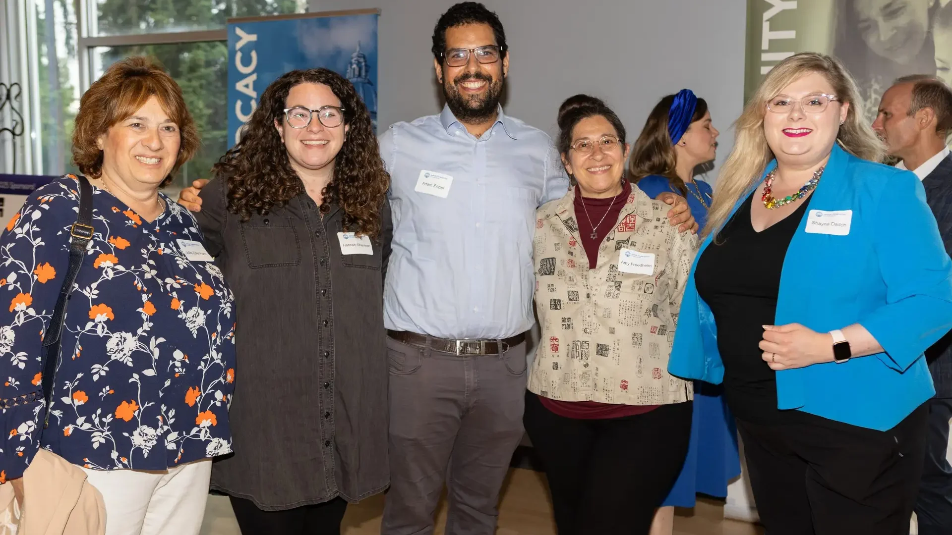 Group of five adults posing together at an event, wearing name tags, with banners in the background.