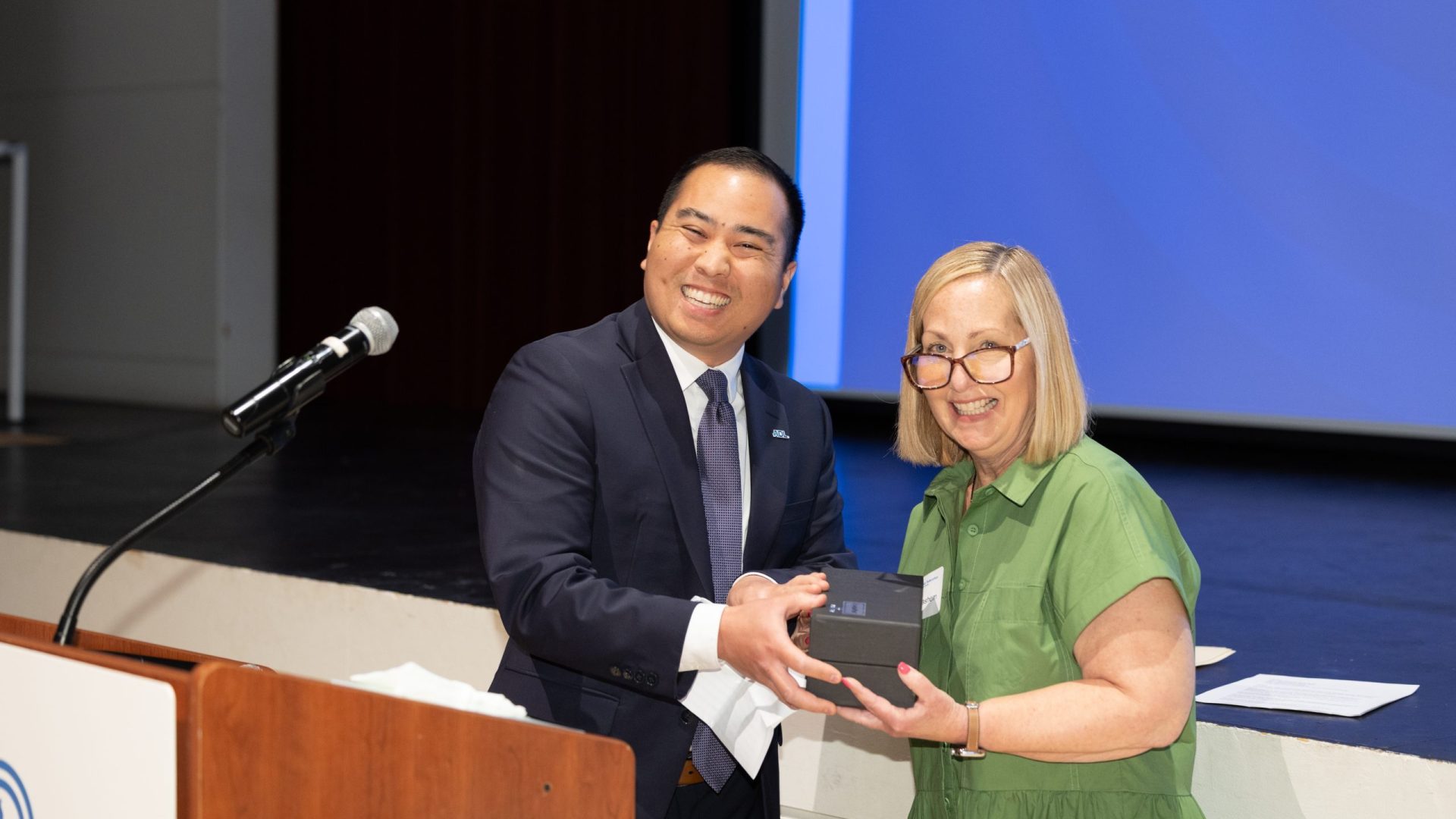 Two presenters on a stage exchange a small box or award while smiling for the camera, with a microphone on a stand to the left and a blue screen in the background.