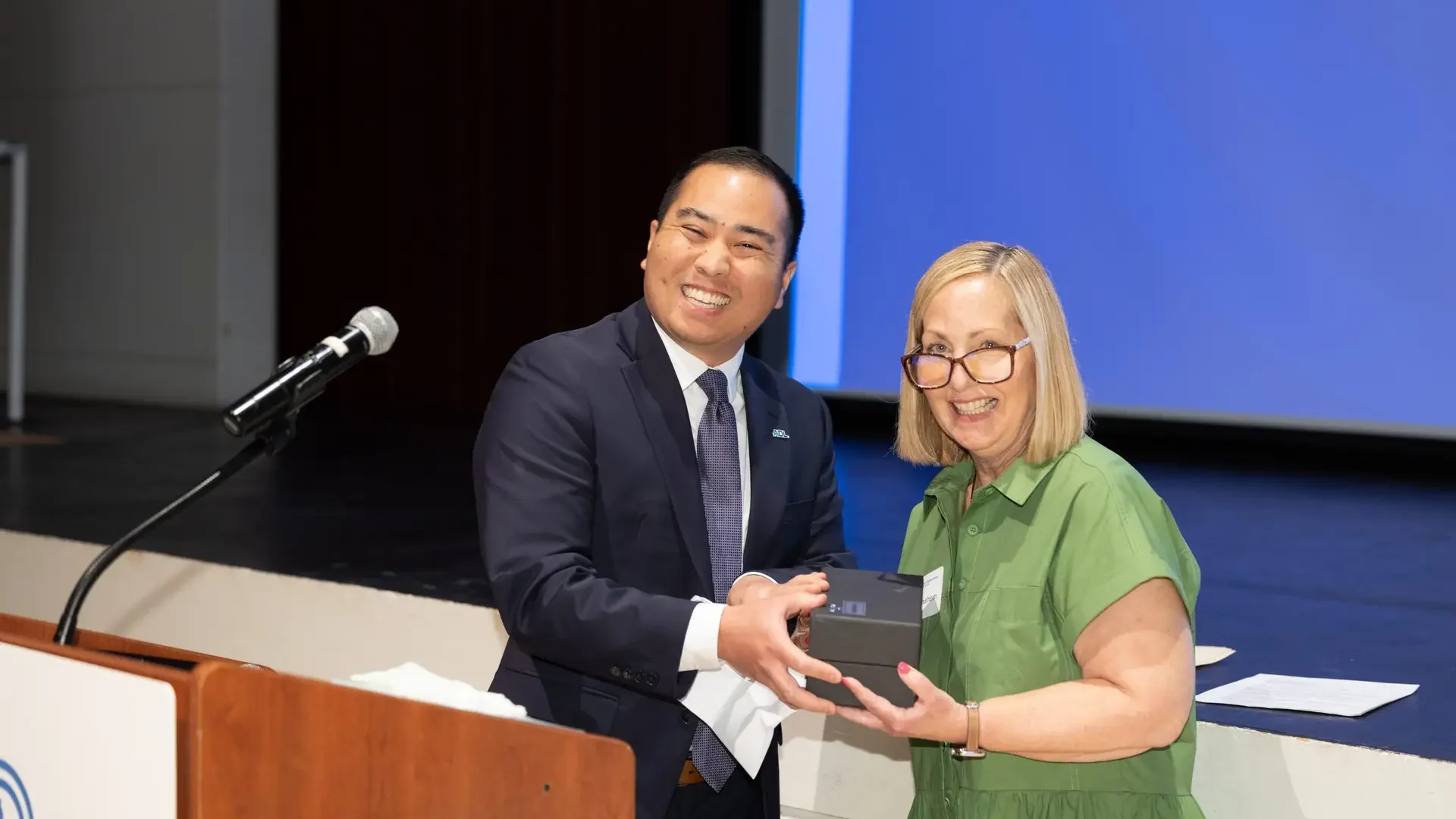 Two presenters on a stage exchange a small box or award while smiling for the camera, with a microphone on a stand to the left and a blue screen in the background.