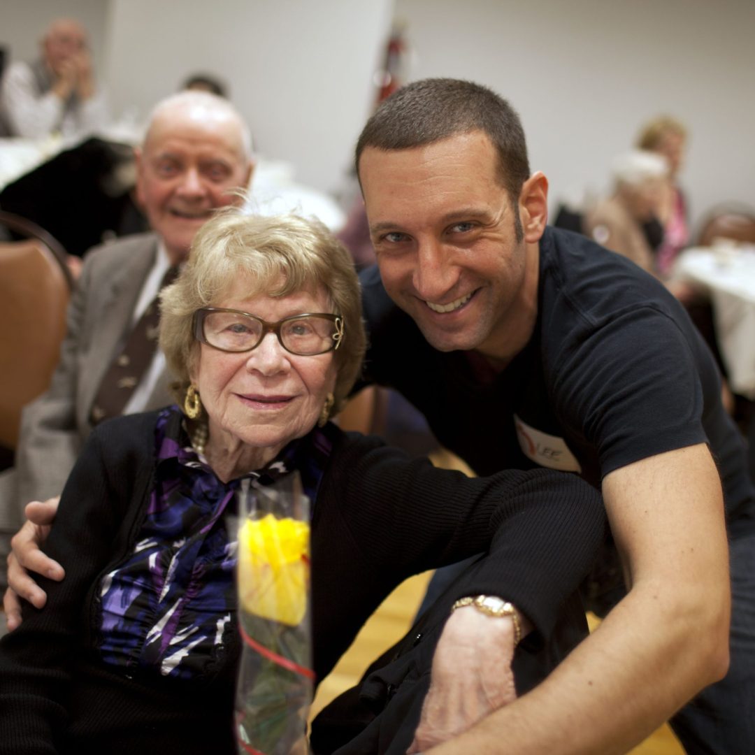 Smiling elderly woman with glasses sits at a table while a smiling man leans in beside her at a social gathering.