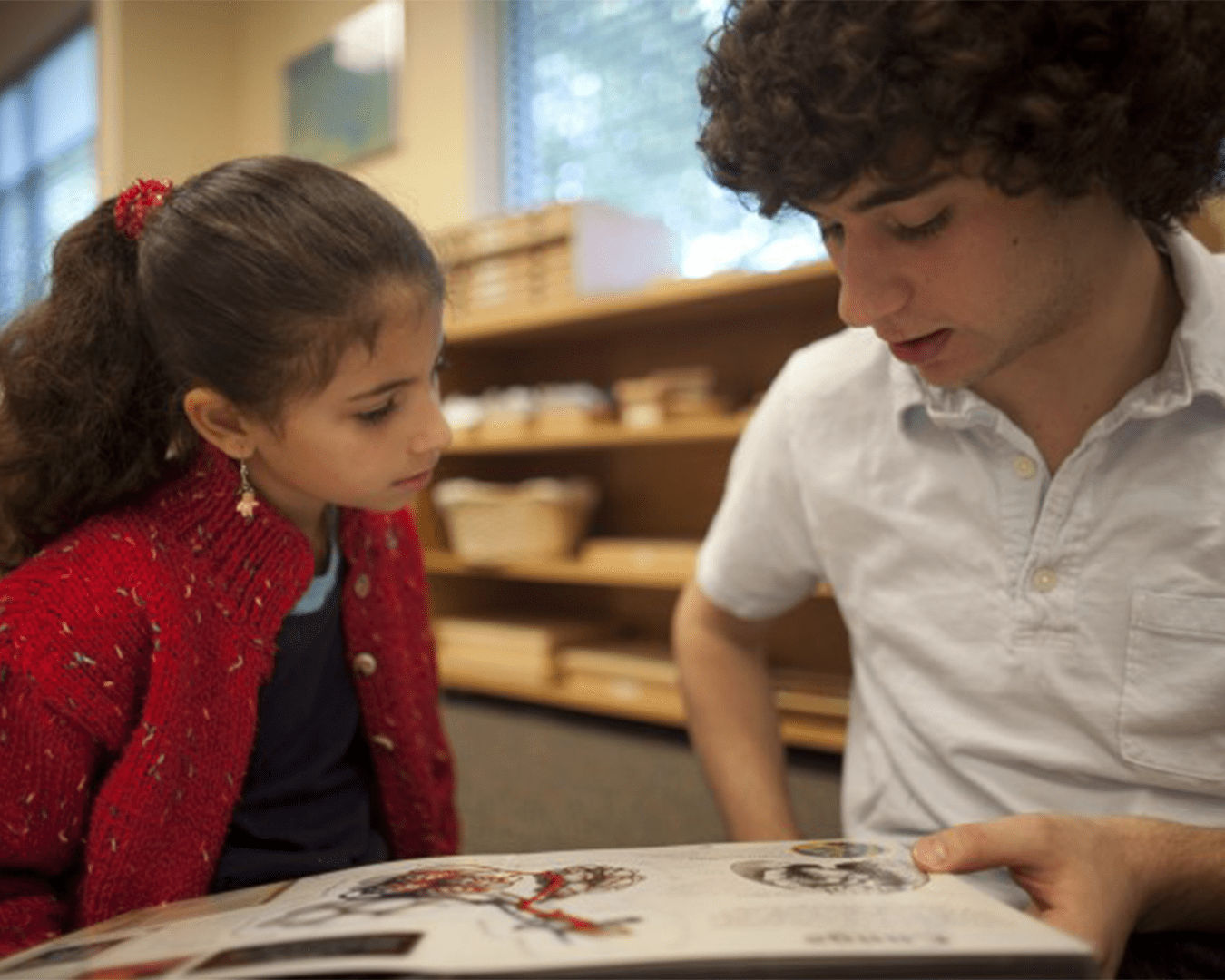 Girl in a red cardigan and a boy in a white shirt read a picture book together at a table in a library, focused on the pages.
