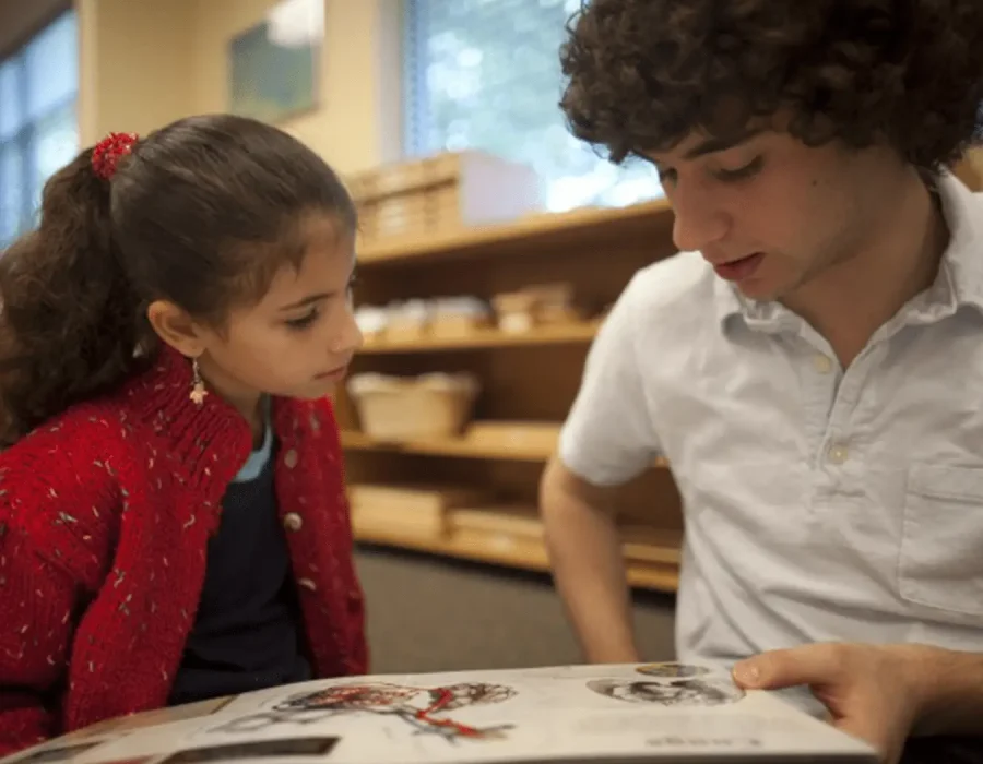 Girl in a red cardigan and a boy in a white shirt read a picture book together at a table in a library, focused on the pages.
