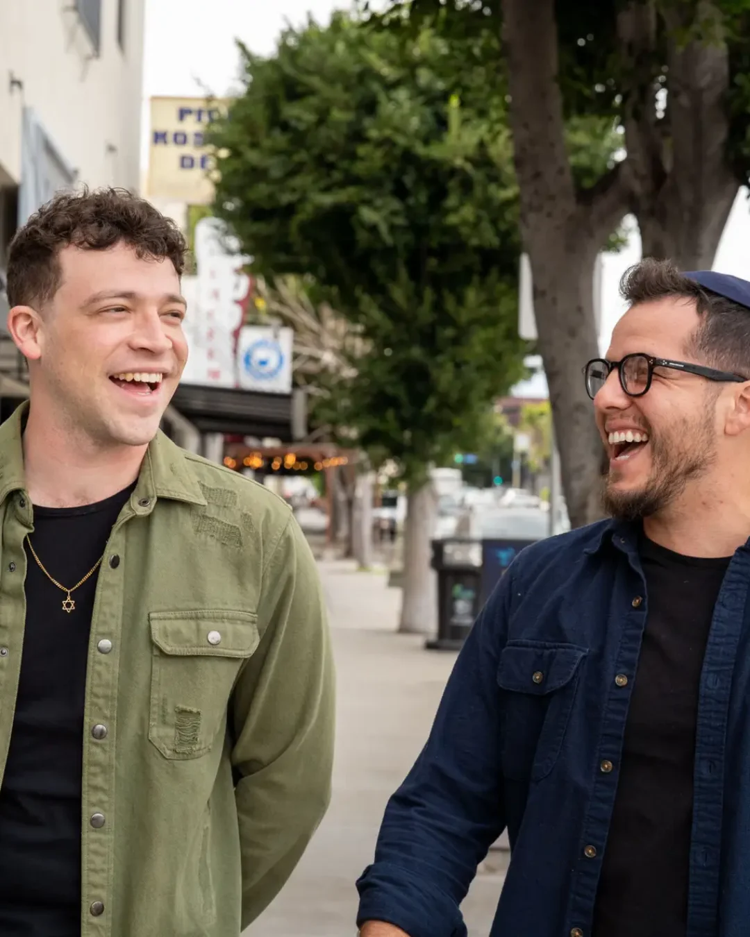 Two men laugh together while walking on a city sidewalk with buildings and trees in the background.