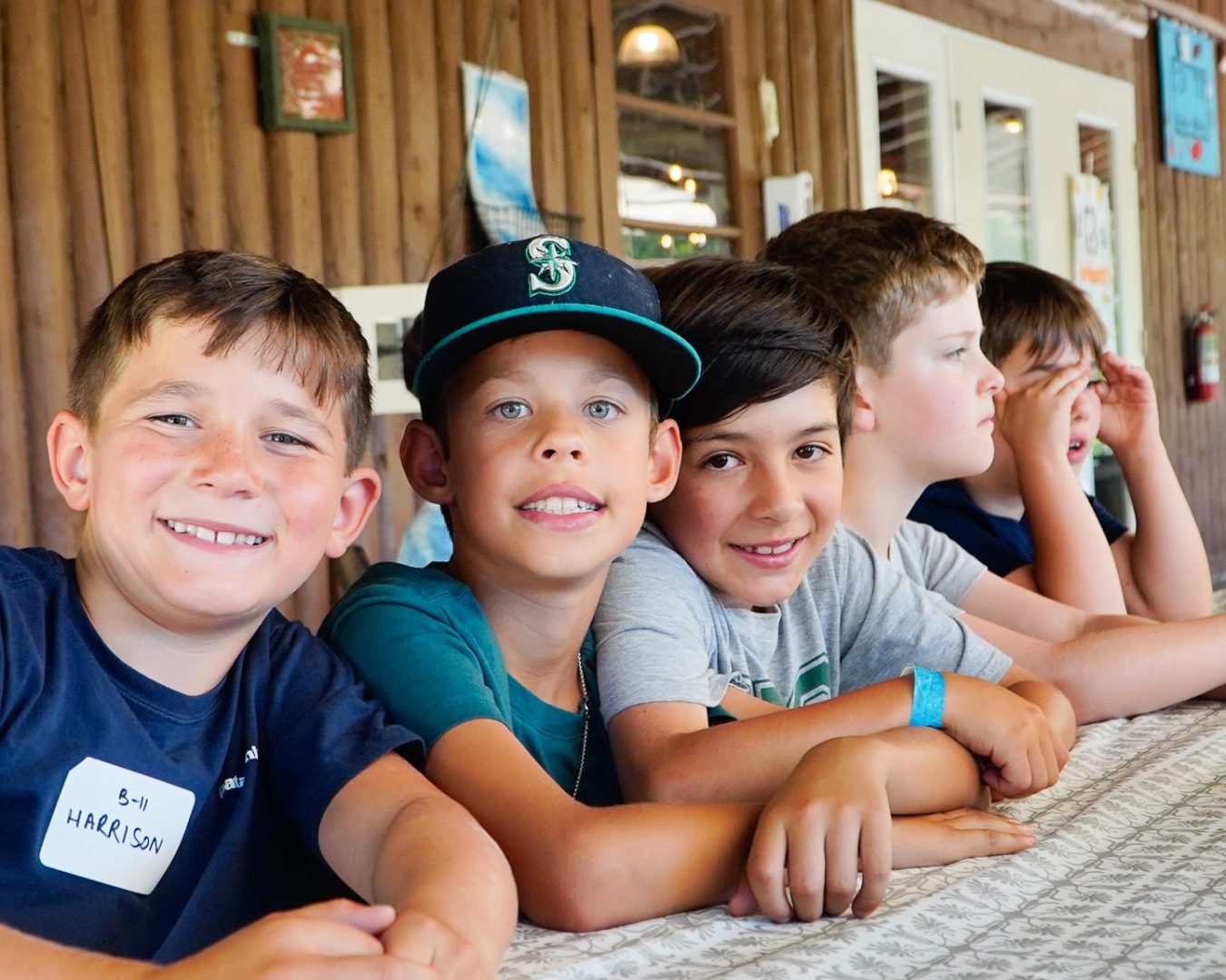 Six kids sit at a long table in a wood-paneled cabin, smiling at the camera.