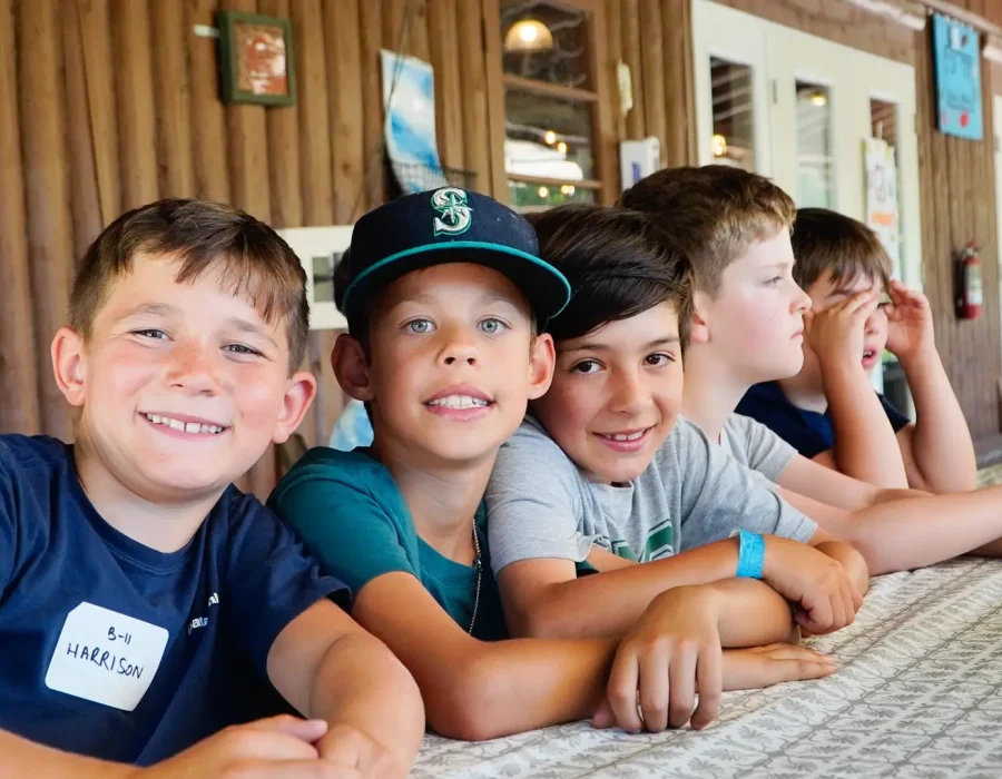 Six kids sit at a long table in a wood-paneled cabin, smiling at the camera.
